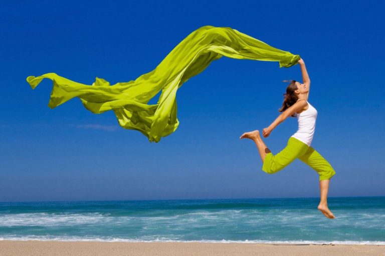 A woman jumping on the beach with a green scarf.